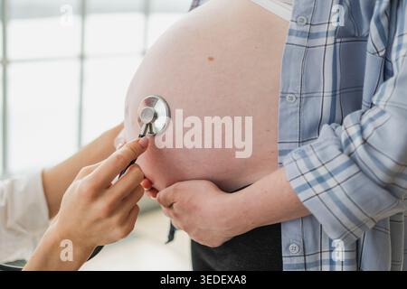 Arzt untersucht schwangere Frau, die ein Stethoskop in der Nähe des großen Bauches hält und den Herzschlag des Babys hört. Gynäkologischer Arzt, der Patientin über Schwangerschaft in der Krankenhauspraxis berät. Besuch bei ärztlicher Untersuchung Stockfoto