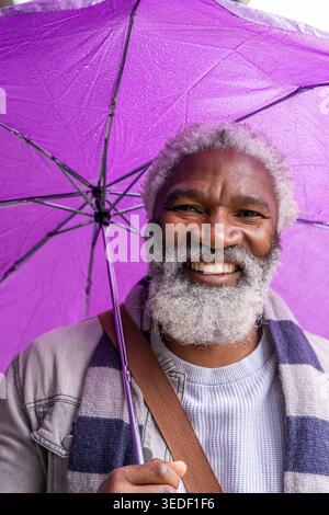 Reifer afroamerikanischer Mann lächelt und hält lila Regenschirm, trägt bei Regen einen Schal Stockfoto