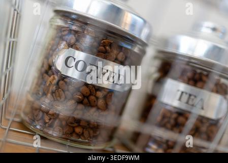 Aus nächster Nähe sehen Sie die Vorratsdosen aus Glas mit der Aufschrift „Coffee“ und „Tea“, die reichhaltige Kaffeebohnen zeigen. Das Bild fängt eine gemütliche Küchenatmosphäre ein, perfekt Stockfoto