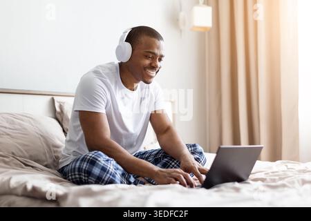 afroamerikaner, der auf dem Bett sitzt und auf einem Laptop im Schlafzimmer schreibt Stockfoto
