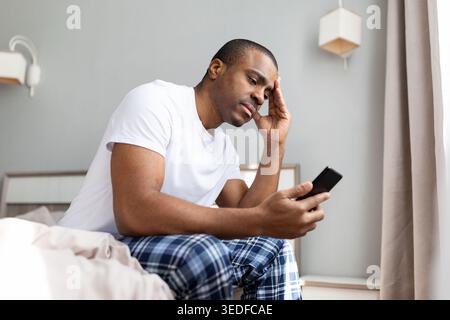 afroamerikanischer Mann, der auf dem Bett sitzt und das Smartphone mit besorgtem Ausdruck anschaut Stockfoto