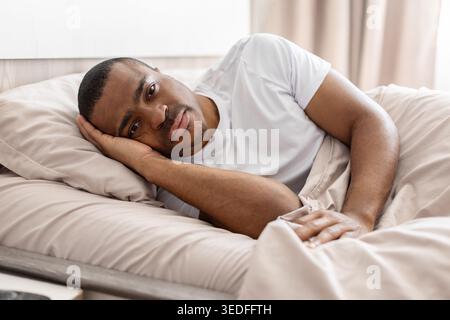 Nachdenklicher afroamerikanischer Mann in weißem T-Shirt, der im Bett auf Kissen liegt Stockfoto