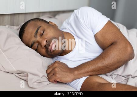 Schwarzer Mann in weißem T-Shirt, der friedlich auf seiner Seite im Bett schläft Stockfoto