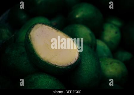 Eine Nahaufnahme einer geschnittenen, rohen grünen Mango, die ihr helles Inneres enthüllt, umgeben von einem Haufen grüner Mangos in Dhaka, Bangladesch. Stockfoto