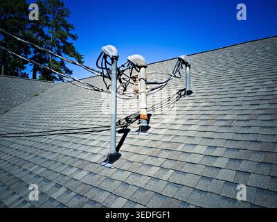 Drei elektrische Versorgungsmasten, die komplexe Verkabelung und elektrische Leitungen unterstützen, erheben sich von einem grauen Asphaltschindeldach unter klarem blauen Himmel. Stockfoto