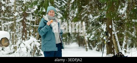 Eine vierzigjährige Frau mit Keks und Silberbecher heißer Schokolade in einem Panorama-Winterwald. Die Dame in blauer Jacke und weißem Schal genießt einen süßen Snack. Horizontales Banner mit ausladender Schneeszene Stockfoto