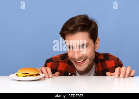 Der Mann sieht Hamburger auf weißem Tisch mit blauem Hintergrund eifrig an Stockfoto