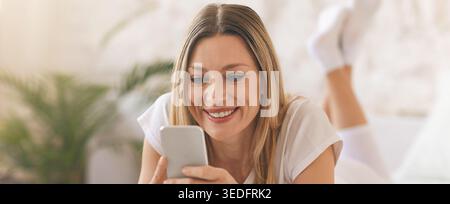 Frau, die ihr Smartphone benutzt, während sie sich auf dem Bett in einem gemütlichen Zimmer mit Pflanzen entspannt Stockfoto