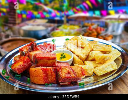 Brasilianische gebratene Schweinerippchen und Maniok (Mandioca) auf einem Teller serviert, mit einem festlichen Juni-Festival (Festa Junina) Hintergrund mit bunten Flaggen und Stockfoto