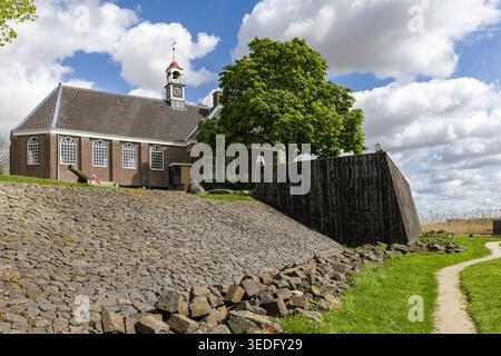 Blick auf die historische Kirche von Schokland mit ihrem Glockenturm und einem großen grünen Baum hinter einem Steindamm unter einem bewölkten Himmel in Schokland, Flevola Stockfoto