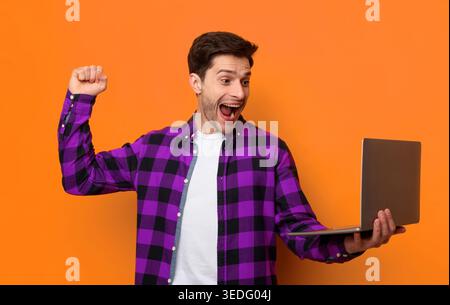 Junger Mann feiert, während er einen Laptop vor einer orangefarbenen Wand hält Stockfoto