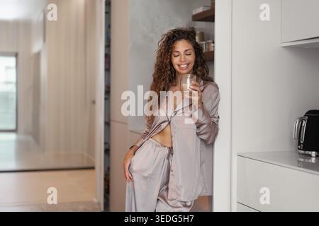Lächelnde Frau mit lockigem Haar, die einen Seidenpyjama trägt, der eine Tasse Kaffee hält, entspannter Morgenmoment in der modernen Küche Stockfoto
