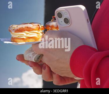 Nahaufnahme von Händen mit veganem Choux-Gebäck mit essbarem Gold, einem Smartphone und aufgerolltem tschechischem Bargeld auf dem Naplavka Street Food Markt. Authentisches Mo Stockfoto