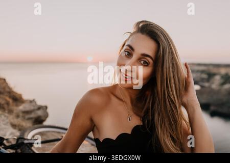 Frau Sonnenuntergang Küste lächelnde junge Frau mit langen Haaren genießt einen friedlichen Abend am Meer, während sie auf einer Klippe posiert Stockfoto