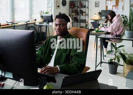 Porträt eines jungen Schwarzen Mannes, der am Desktop-Computer arbeitet, während er Kopfhörer in einem modernen Büro trägt, Frau, die im Hintergrund arbeitet. Ideal für Teamarbeit, Produktivität und Startup-Marketing Stockfoto