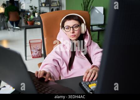Junge Erwachsene kaukasische Frau mit Kopfhörern arbeitet am Computer im Büro, verwaltet digitale Aufgaben und Multitasking. Nützlich für Fernarbeit, Produktivität und technische Karriereinhalte Stockfoto