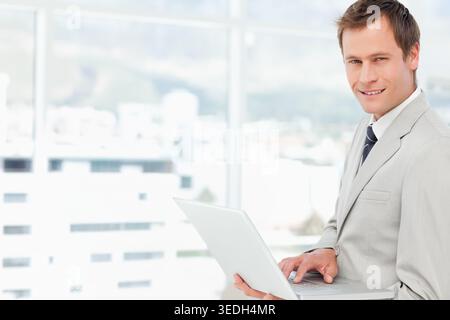 Männlicher Mann, der im Büro an den Fenstern steht, Anzug und Krawatte mit silbernem Laptop hält, Kopierraum. Professionell, Geschäftsmann, leitend, städtisch, s Stockfoto