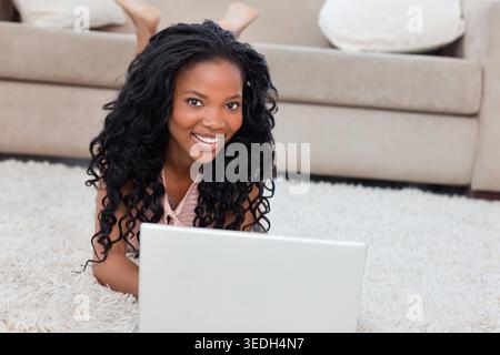 Afroamerikanerin lächelt, während sie zu Hause auf weißem Teppich liegt, indem sie einen Laptop benutzt. Arbeit, Fernbedienung, Gerät, Wohnzimmer, Sofa, Teppich, gemütlich Stockfoto