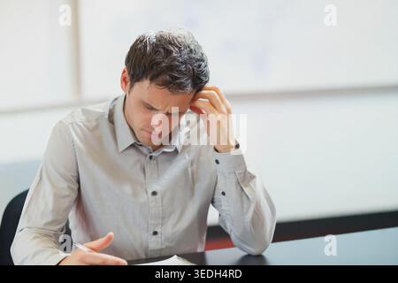 Mann im mittleren Erwachsenenalter mit grauem Hemd, der am Schreibtisch sitzt und den Stift in einem Spiralheft hält. Fokus, Kontemplation, Solo, professionell, modern, min Stockfoto