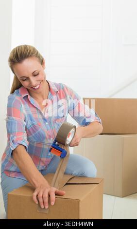 Frau im mittleren Erwachsenenalter kniend, versiegelnde Umzugskarton mit Klebeband zu Hause mit pastellfarbenem kariertem Hemd. Verpackung, Umzug, Sonnenlicht, Innenraum, organisiert, Stockfoto