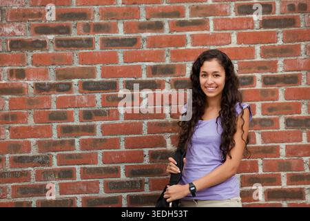 Erwachsene Frau, die direkt an der roten Ziegelwand steht, schwarze Tasche hält, Digitaluhr zeigt, Kopierraum. Person, Urban, Lifestyle, Mode, Umhängetasche, Zeit Stockfoto