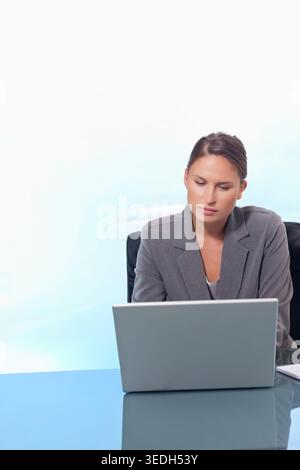 Büroangestellte mit grauem Blazer, die am Glasschreibtisch sitzt, mit silbernem Laptop und Notebook. Frau, professionell, minimalistisch, modern, Arbeitsplatz, Stockfoto