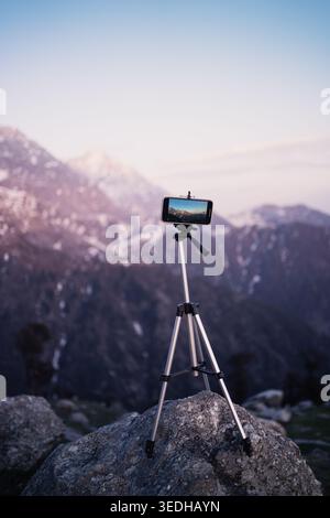 Ein auf einem Stativ montiertes Mobiltelefon steht auf einem Felsen mit Blick auf die verschneiten Gipfel in Triund, Mcleodganj, Himachal Pradesh, Indien. Stockfoto
