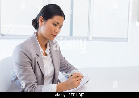 Asiatische Frau, die am weißen Schreibtisch sitzt, einen hellgrauen Blazer trägt und mit Stift auf Notizblock schreibt. Minimalistisch, sauber, modern, professionell, am Arbeitsplatz, Stockfoto