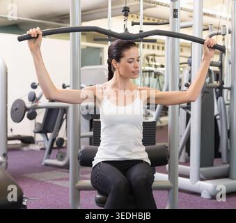 Frau mit weißem Tank und schwarzer Leggings, die an der Pulldown-Maschine des Fitnessstudios eine geschwungene Pulldown-Bar zieht. Fitnesscenter, Kraft, Training, Workout, Ausrüstung Stockfoto