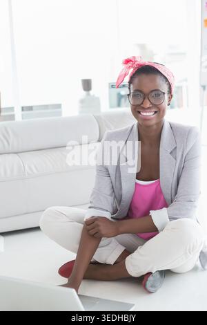 Afroamerikanerin mit grauem Blazer, rosa Schal sitzt zu Hause auf dem Boden und arbeitet am Laptop. Computer, minimalistisch, Tageslicht, Wohnzimmer, Fernbedienung Stockfoto
