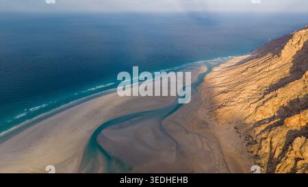 Blick aus der Vogelperspektive auf einen Wüstenstrand an der Küste mit einem gewundenen Gezeitenkanal, der neben sonnendurchfluteten felsigen Klippen unter trübem Himmel auf das blaue Meer trifft. Detwah Lagune, Also Stockfoto