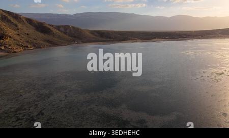 Aus der Vogelperspektive auf eine ruhige Küstenlagune oder Bucht bei Sonnenuntergang mit Hügeln und Bergen im Hintergrund, sanftes goldenes Licht reflektiert auf flachem Wasser in der Nähe eines Stockfoto