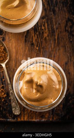 Cremiger, gepeitschter Dalgona-Kaffee-Dessert in Glasgläsern auf rustikalem Holztisch mit Löffel und Kaffeesatz Stockfoto