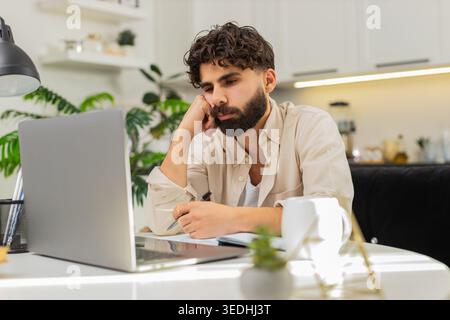 Indischer Mann sitzt zu Hause mit Laptop fühlt sich sehr faul abgelenkt hält Kopf nahe einer Tasse Kaffee und Notizen. Ein arabischer Kerl, der am Tisch sitzt, starrt auf Bildschirme, arbeitet an Aufgaben, fühlt sich an, dass wenig Energie Pause will. Stockfoto