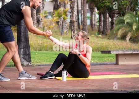 Die reife Frau sitzt auf der Trainingsmatte und erhält Unterstützung vom Personal Trainer während des Workouts im Freien in einem tropischen Park. Gesundes Leben Stockfoto