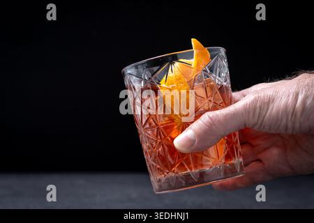 Nahaufnahme einer Hand mit einem klassischen altmodischen Whiskey-Cocktail, serviert in einem Kristallglas mit Eis- und Orangenschalen-Garnitur vor einem dunklen Hintergrund Stockfoto