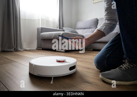 Smart-Home-Konzept mit einer Person, die ein Mobiltelefon benutzt, um einen Roboter-Staubsauger auf einem Holzboden in einem modernen Wohnzimmer zu steuern. Heimautomatisierung, Stockfoto