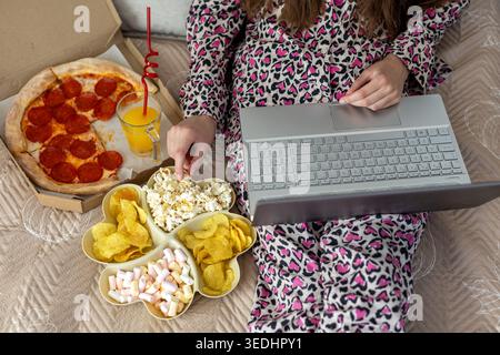 Frau, die einen Laptop im Bett mit Snacks beobachtet. Gemütliches Lifestyle-Foto von einer Frau im Pyjama mit einem Laptop im Bett mit Pizza, Saft, Popcorn, Chips Stockfoto