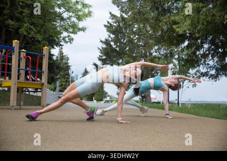 Sportliche zwei Frauen im Sportoutfit, die morgens trainieren und sich auf dem Straßensportplatz Strecken. Fitness-Workout auf dem Sportplatz. Ausbildung bei einem Inruktor in der Natur Stockfoto