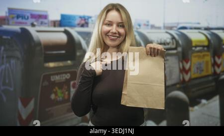 Eine blonde, lächelnde Frau, die eine braune Papiertüte hält, zeigt auf der städtischen Straße mit dem Finger auf die Brust, indem sie Abfalleimer recycelt; Selbstvertrauen stärkt. Stockfoto