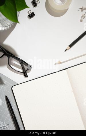 Rahmen aus offenem gepunktetem Notizbuch, schwarzem Stift, Brille, Büroklammern, Klebeband, Bleistifte und Schneidematte auf weißem Schreibtischhintergrund. Kopierraum Stockfoto