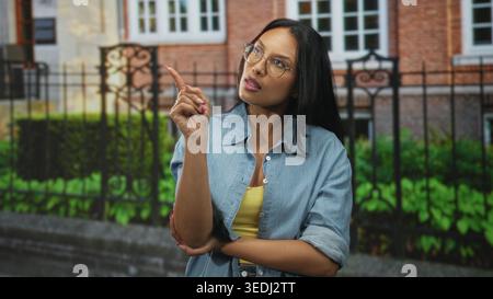 Frau zeigt mit erhobener Hand und gefaltetem Arm vor das Gebäude, trägt Brille und Jeansshirt; Verwirrung. Stockfoto