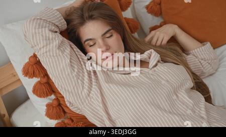 Frau, die mit erhobenem Arm und geschlossenen Augen auf dem Bett liegt, sich an einem Kissen mit Quasten im Gebäude auflegt, gestreiftes Pyjamaoberteil und entspannter Haltung trägt; Ser Stockfoto