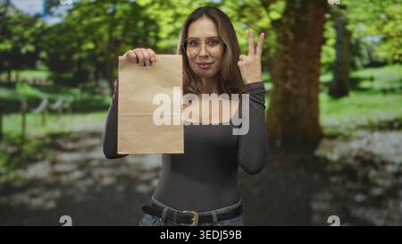 Frau mit brauner Papiertüte und Zeigefinger im Wald mit Brille und Jeans; Selbstvertrauen-Abgabe-Promotion. Stockfoto
