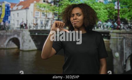 Frau zeigt auf einer amsterdamer Straße an einer Kanalbrücke mit Fahrrädern und Bäumen die Daumen nach unten; Kritik an der Missbilligung. Stockfoto