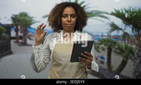 Junge afroamerikanische Bäckerin in Schürze, die Tablette hält und Finger auf der Straßenpromenade am Meer mit Palmen drückt; Präzision Stockfoto