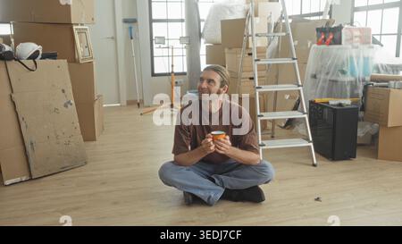 Mann saß im Kreuz und hielt mit den Händen einen orangen Kaffeetasse, blickte zwischen verpackten Kisten, Leiter, Schutzhelm und Werkzeugen in einem Gebäude auf Stockfoto