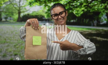 Frau in gestreifter Bluse und Brille zeigt den Finger auf die Papiertüte im Park, während sie in die Kamera lächelt; Markenwerbung glücklich. Stockfoto