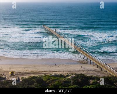 Ein Blick aus der Vogelperspektive auf einen Sandsteg, der sich in das blaue Meer an der Gold Coast erstreckt, mit einem Rettungsschirm-Turm am Strand. Stockfoto