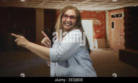 Die Frau zeigt mit dem Finger und lächelt mit den Händen in einem unvollendeten Innenraum des Gebäudes, trägt rote Brille und lässiges Hemd; verspielte Freude. Stockfoto